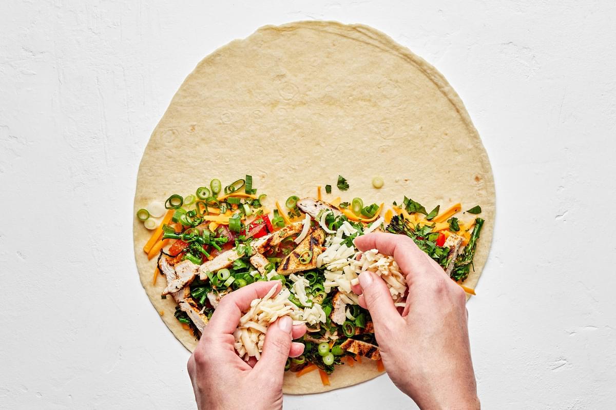 2 hands filling a flour tortilla with cheese, grilled onion, peppers, broccolini, chicken, cilantro and green onions