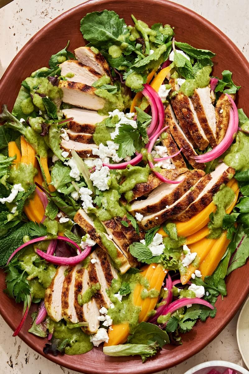 salad topped with grilled curry chicken, mango slices, pickled onion, goat cheese, cilantro, mint and herb vinaigrette