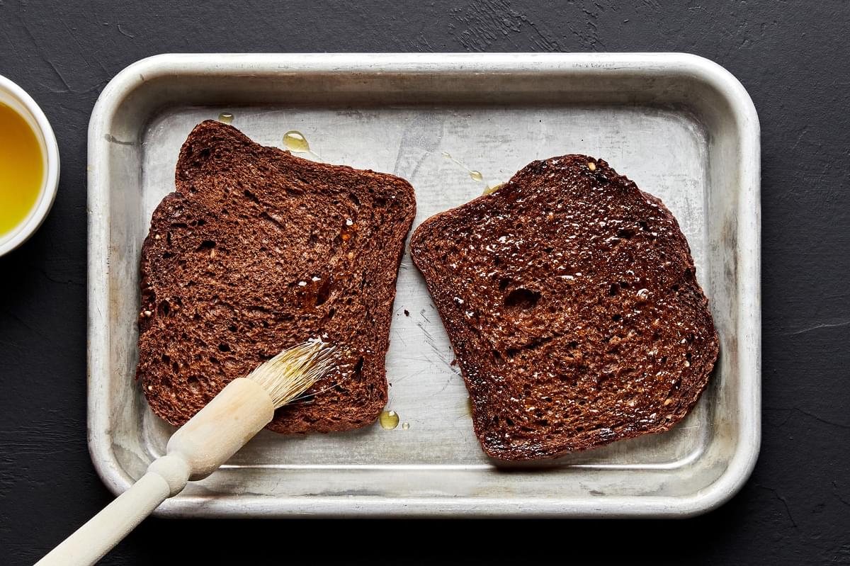 2 slices rye bread on a baking sheet being brushed with olive oil