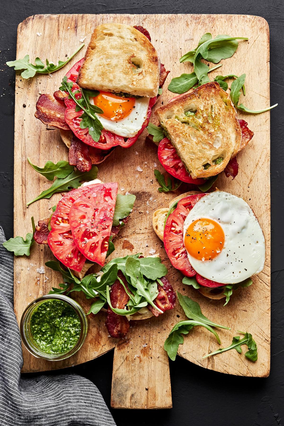 heirloom BLT sandwiches with pesto and fried eggs being assembled on a cutting board
