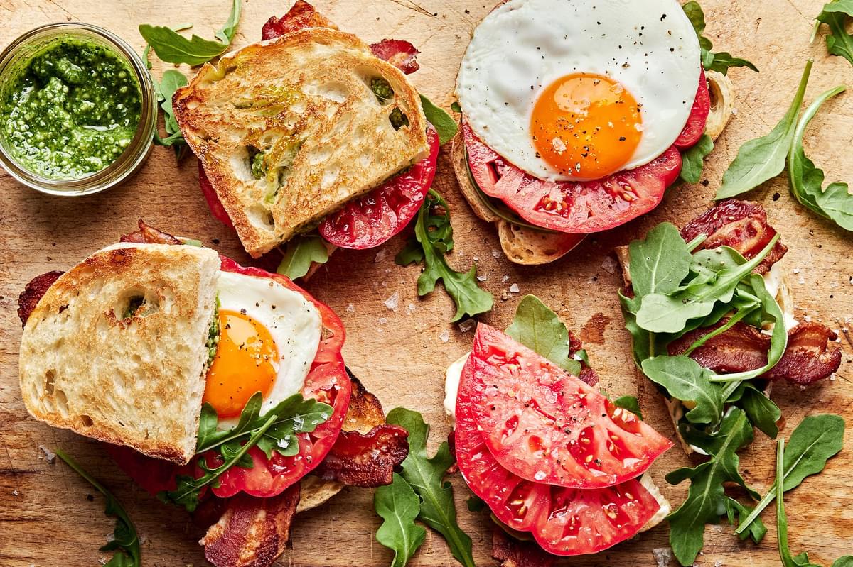 heirloom tomato BLT sandwiches with pesto and fried eggs being assembled on a cutting board