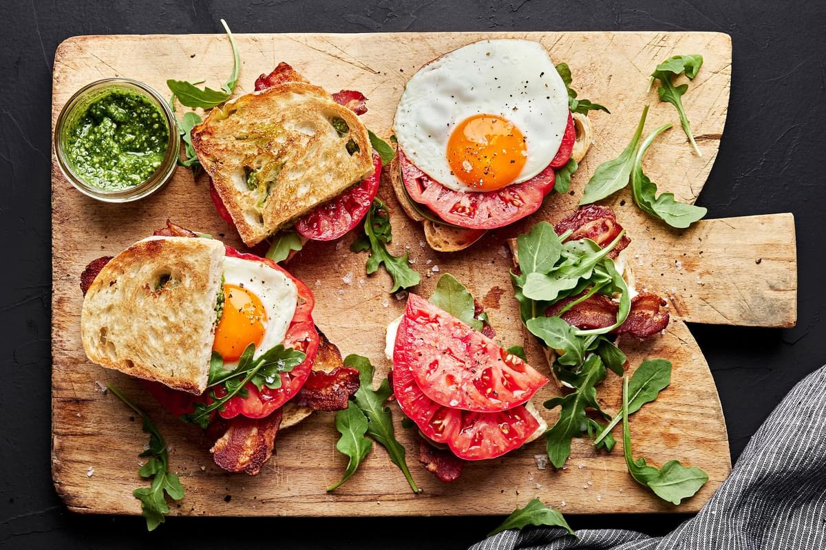 bread being topped with mayo, pesto, bacon, arugula, tomato and a fried egg