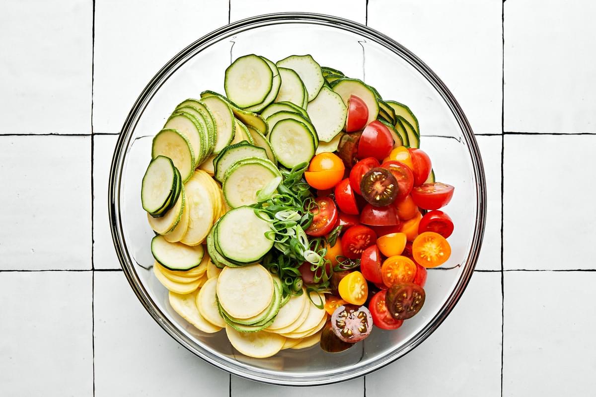 zucchini, squash, tomatoes, and green onions being tossed together in a large bowl