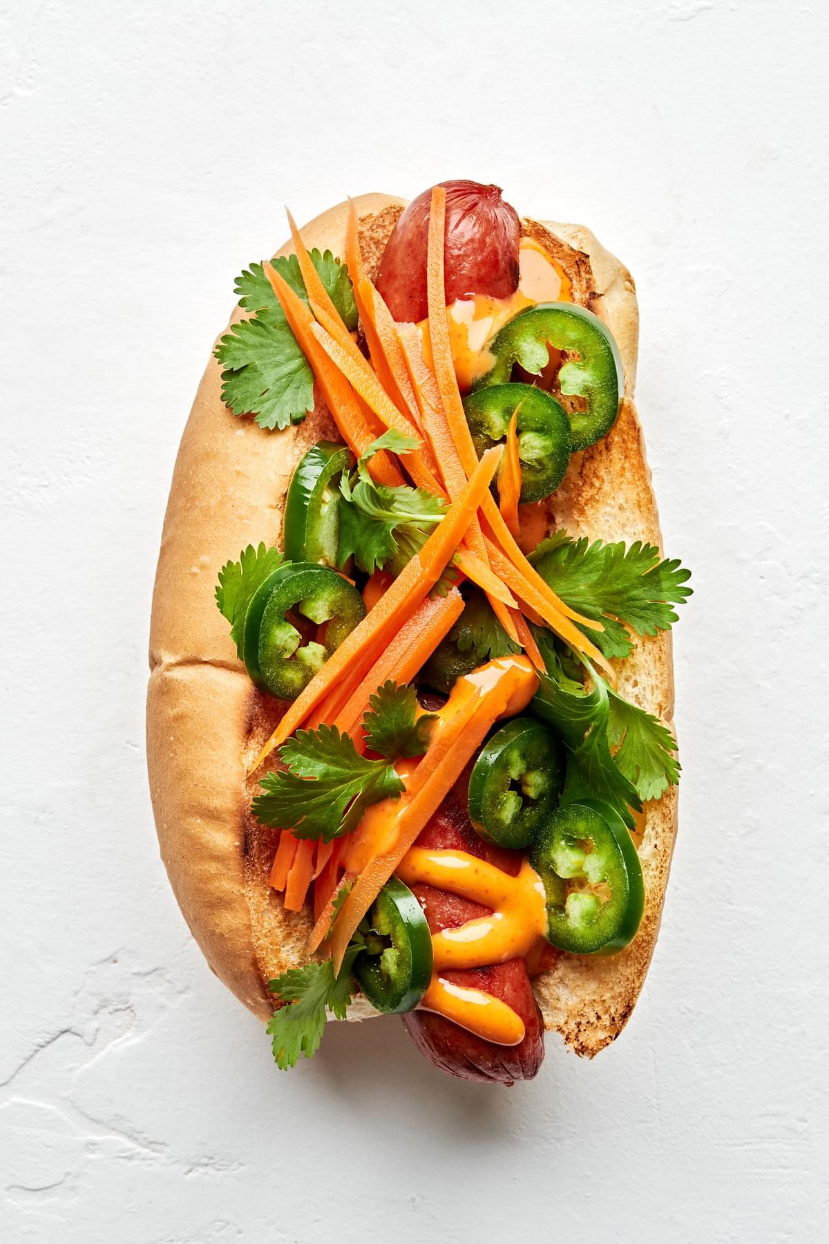 A banh mi hot dog topped with Sriracha mayo, jalapeño, pickled carrots, and cilantro.