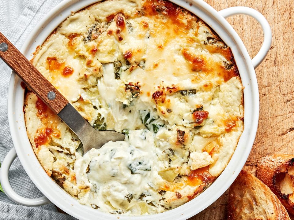 hot spinach and artichoke dip in a baking dish being scooped with a spoon surrounded by toasted crostini for serving