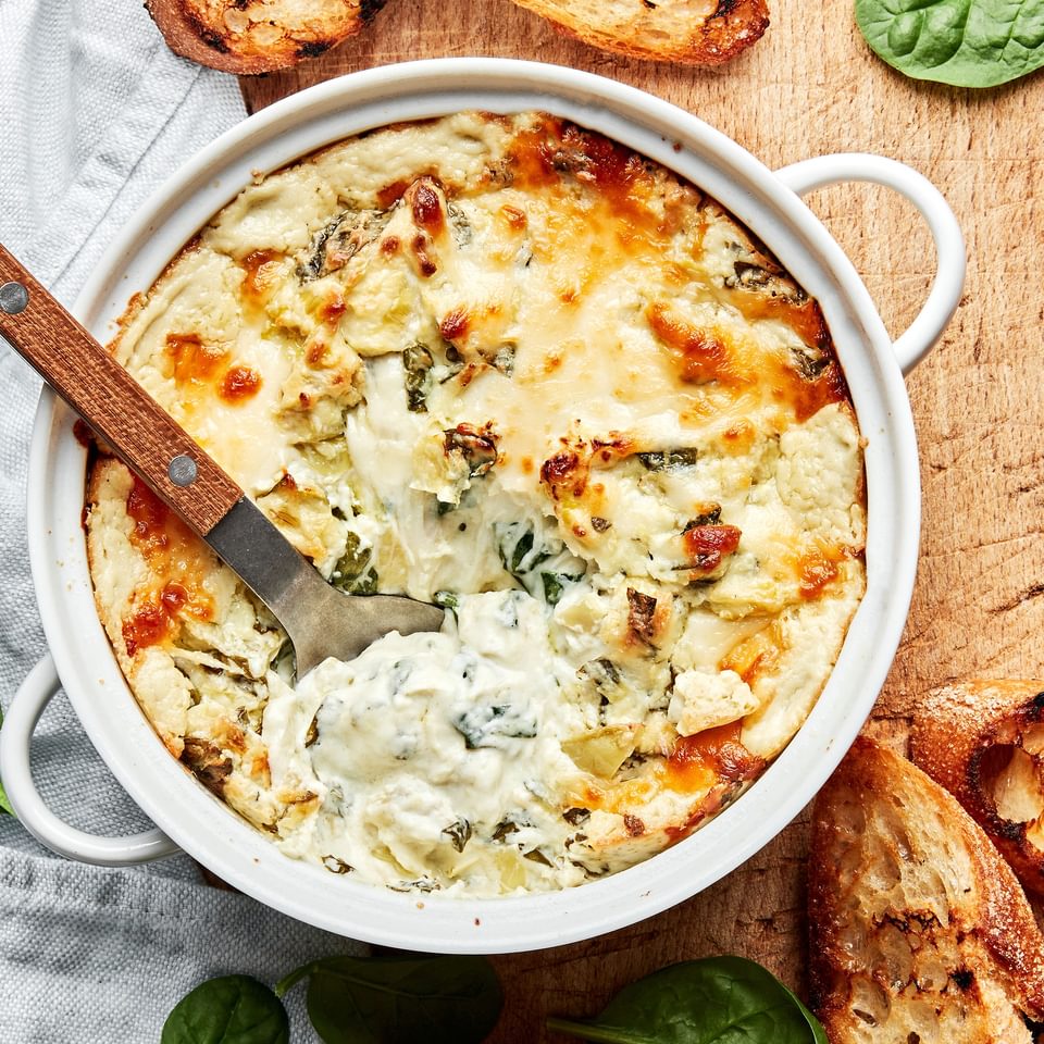 hot spinach and artichoke dip in a baking dish being scooped with a spoon surrounded by toasted crostini for serving