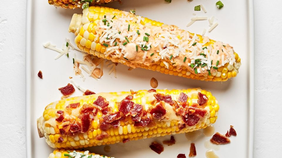 4 grilled corn on the cob with different toppings: maple bourbon butter, chipotle mayo, spicy cotija cheese, & blue cheese