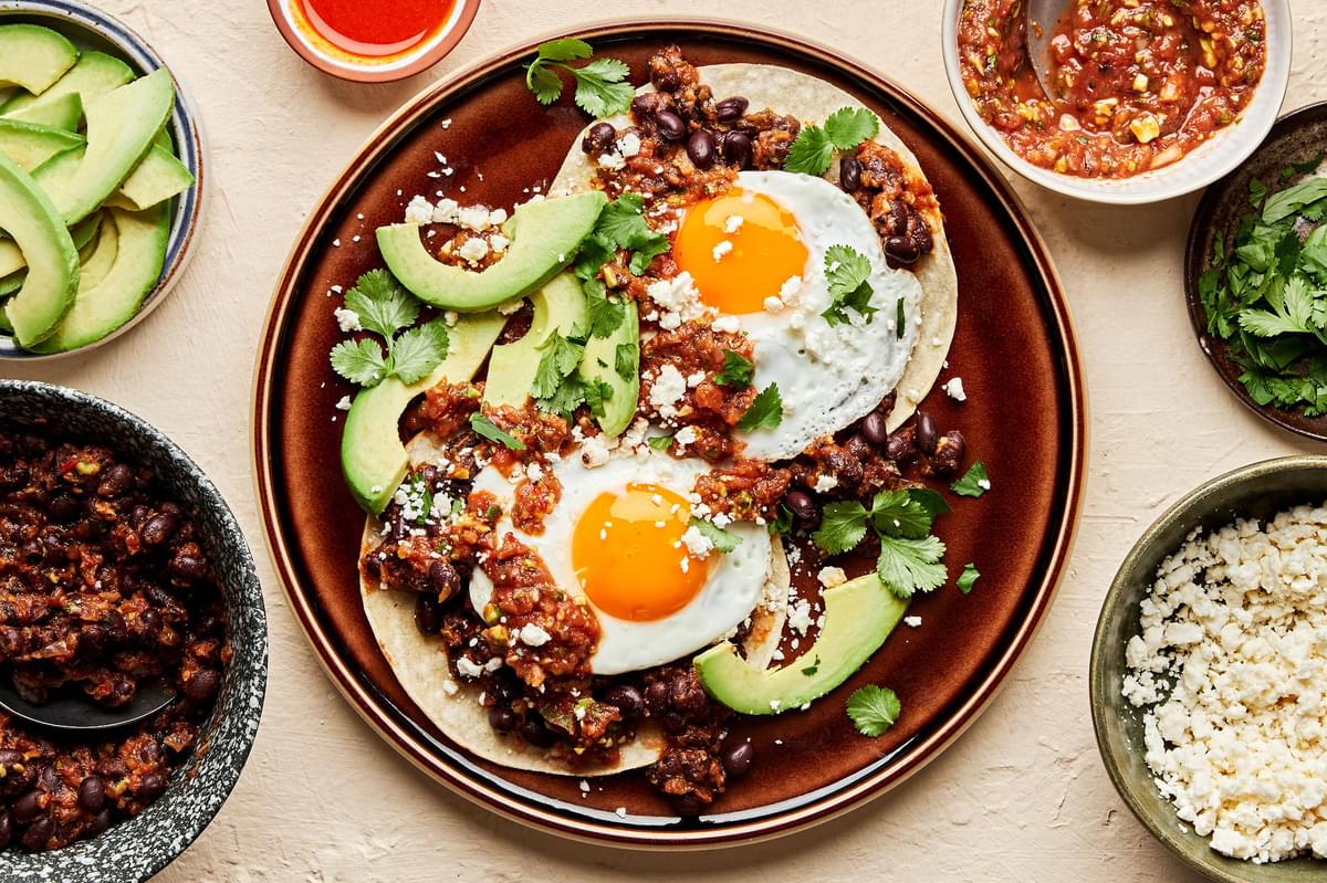 a plate of homemade huevos rancheros surrounded by bowls of toppings: salsa, avocado, cilantro and cotija