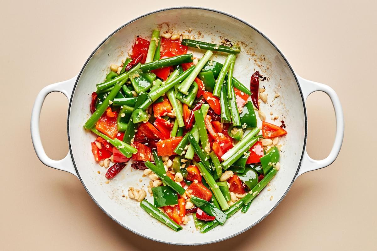 bell peppers, green onions, peanuts, ginger, garlic, and chili peppers being cooked in sesame oil in a skillet
