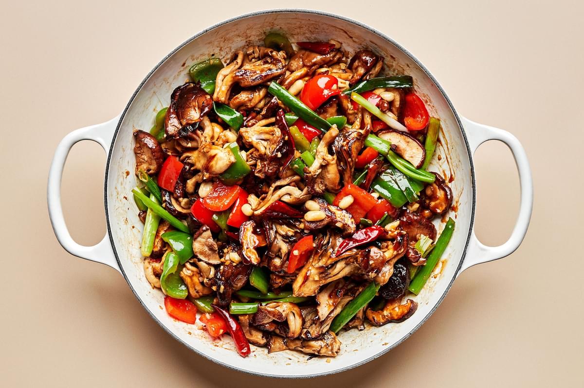 mushrooms, bell peppers, green onions, peanuts, ginger, garlic, and chili peppers being cooked in sesame oil in a skillet