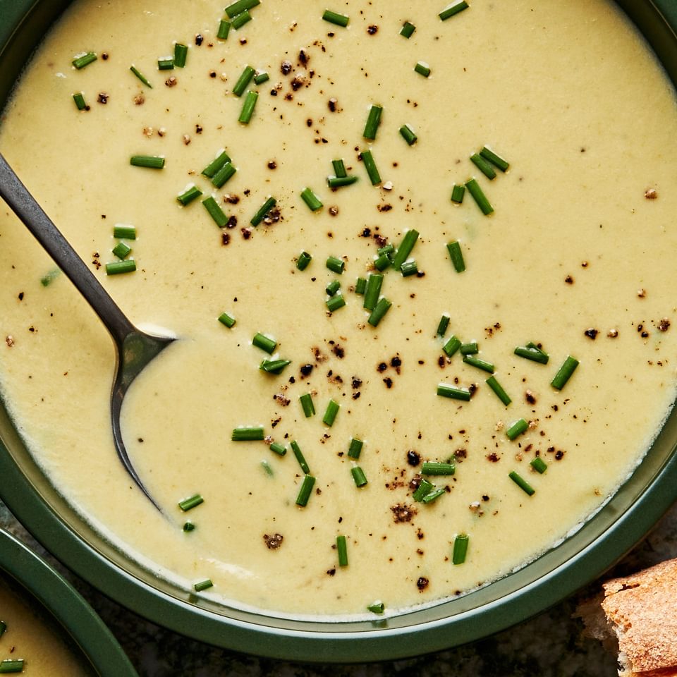 Bowl of creamy leek soup topped with fresh chives and a spoon in it surrounded by crusty bread slices.