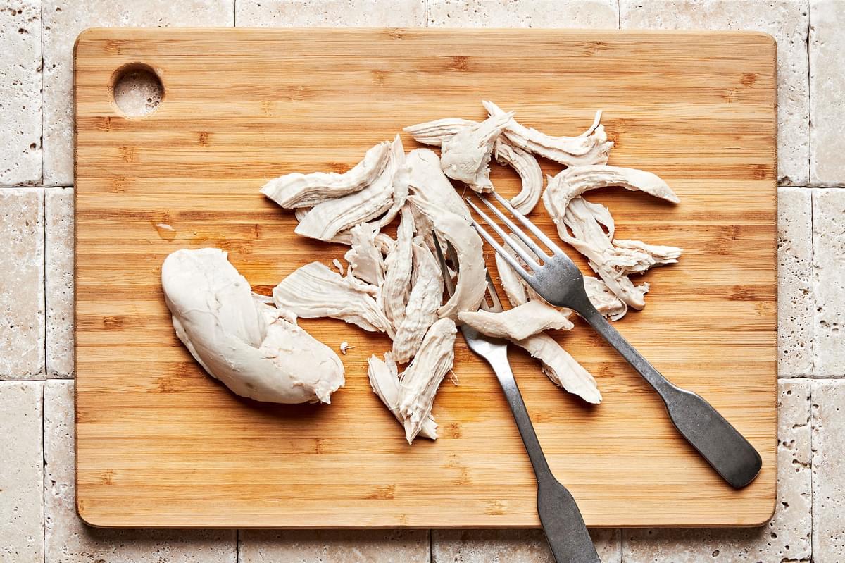 cooked chicken breast being shredded with two forks on a cutting board