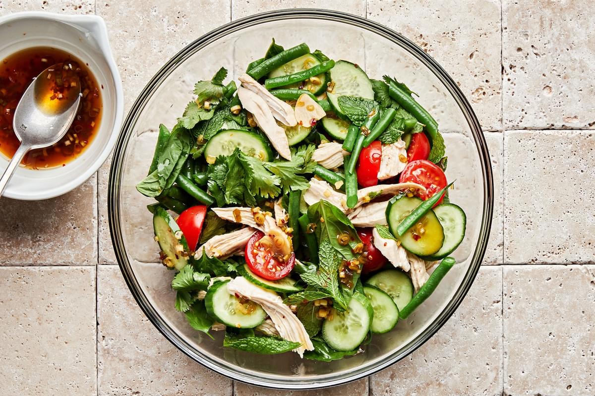 shredded chicken, cilantro, mint, basil, tomatoes, cucumbers to and green beans in a bowl next to a small bowl of dressing