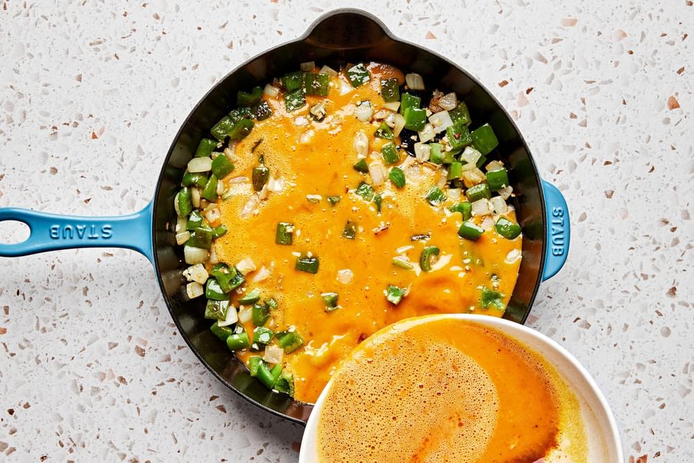 seasoned eggs being poured over bell pepper, onion and garlic in a skillet