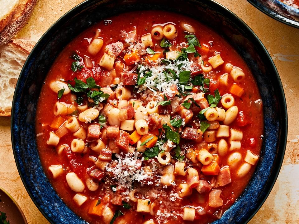 a bowl of homemade pasta e fagioli sprinkled with parsley and parmesan surrounded by slices of crusty bread