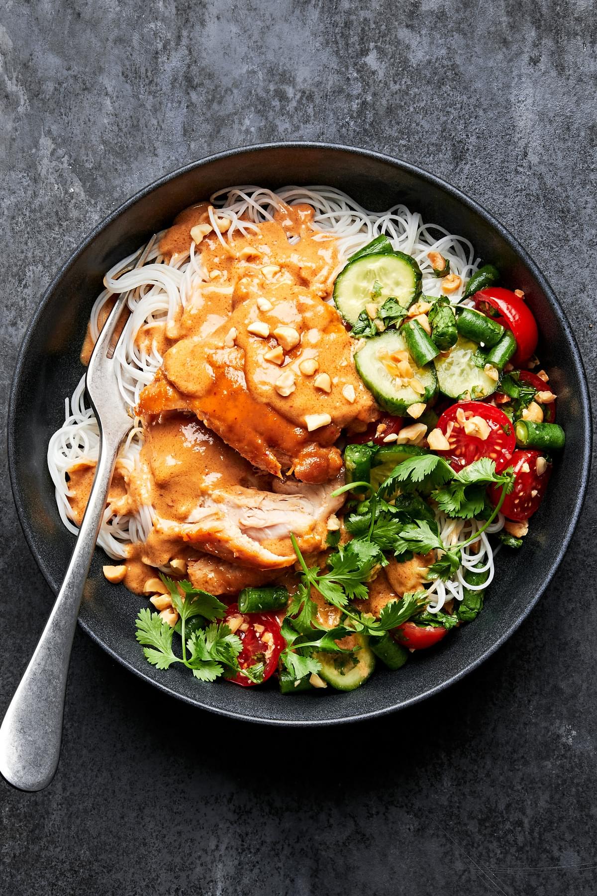 a peanut chicken noodle bowl made with rice noodles topped with chicken, peanut sauce, green bean & tomato salad & cilantro