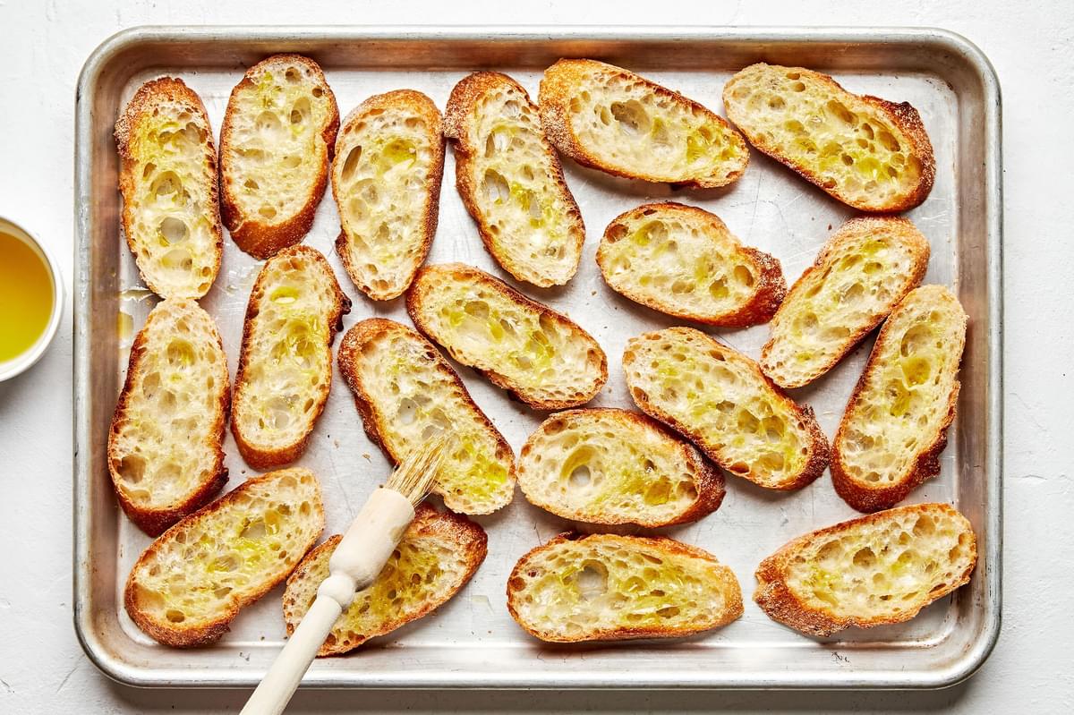 baguette slices on a baking sheet being brushed with olive oil