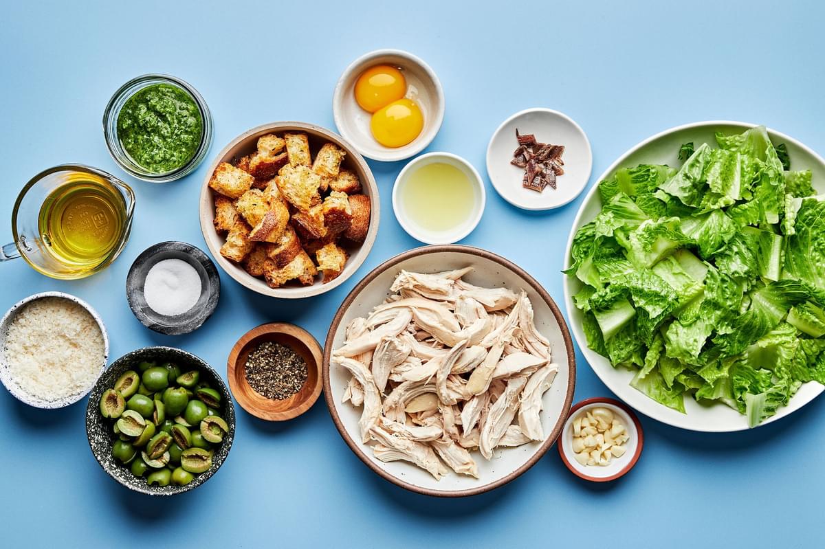 Romaine, shredded chicken, croutons, castelvetrano olives and ingredients to make pesto caesar salad in prep bowls