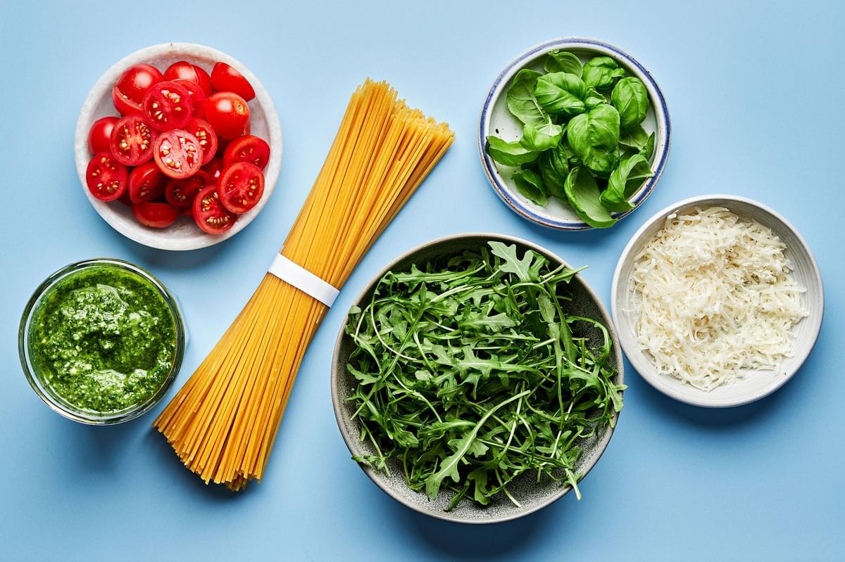 spaghetti, pesto, cherry tomatoes, arugula, basil and parmesan in prep bowls to make pesto spaghetti
