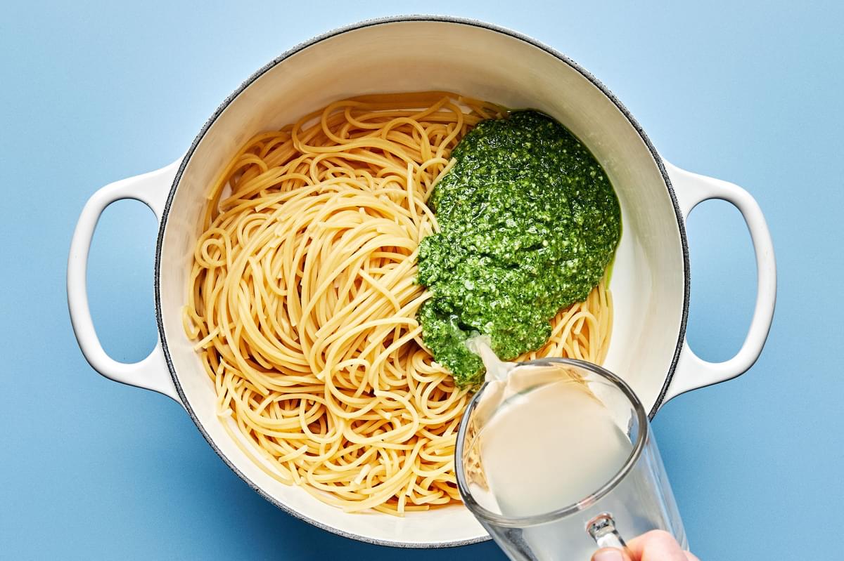 pesto and reserved pasta water being tossed with spaghetti noodles in a pot