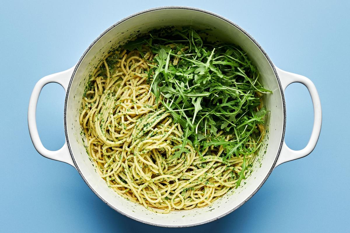 arugula being tossed with pesto and spaghetti noodles in a pot
