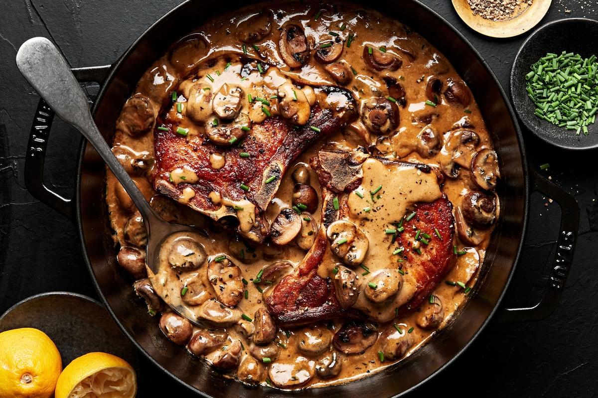 2 bone in pork chops in a skillet filled with creamy mushroom sauce with lemons on the side and a serving spoon