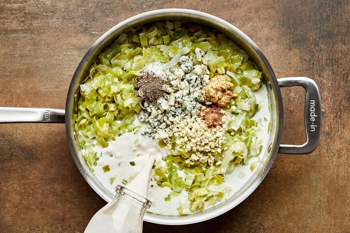 cream being poured into a skillet with leeks, butter, milk, blue cheese, garlic, mustard, nutmeg, salt, and pepper.
