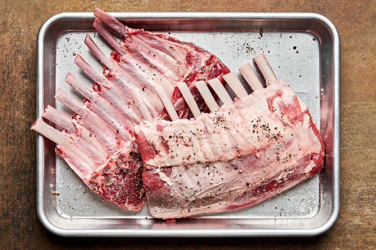 a rack of lamb being seasoned with salt and pepper on a baking sheet