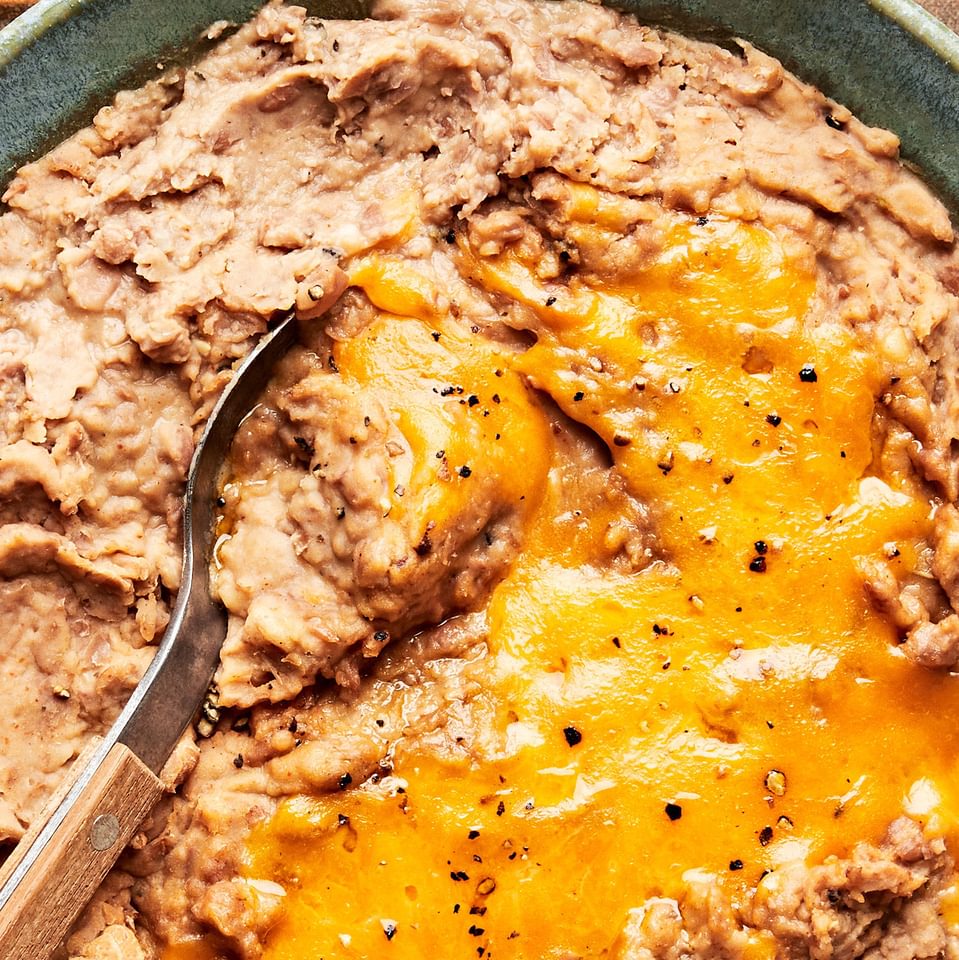 homemade refried beans topped with melted cheddar cheese in a bowl with a spoon