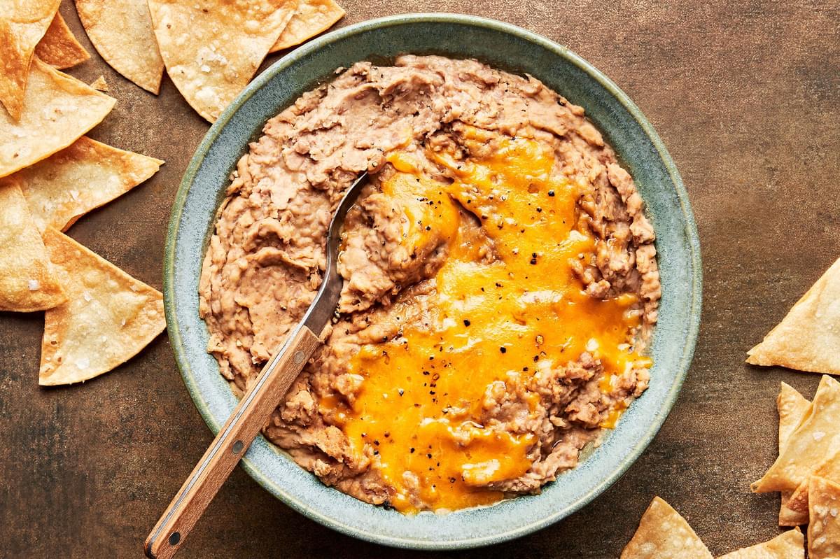 homemade refried beans topped with melted cheddar cheese in a bowl with a spoon surrounded by tortilla chips