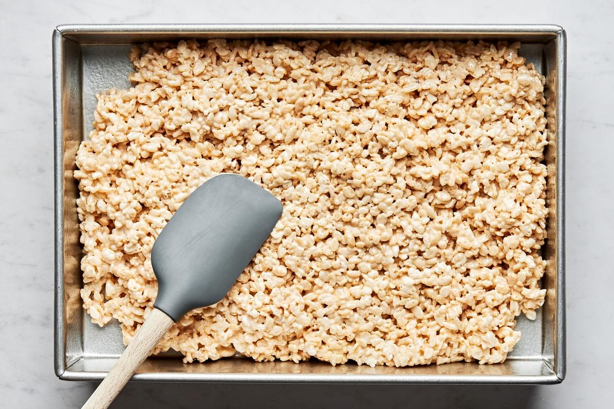 homemade rice crispy treats being spread into a greased 9x13 baking sheet with a spatula
