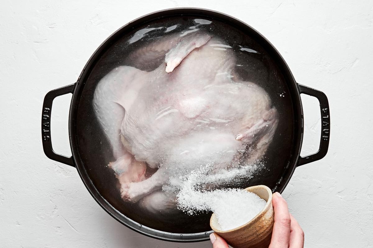a raw spatchcocked turkey in a large pot of water with salt being poured in for brining the turkey