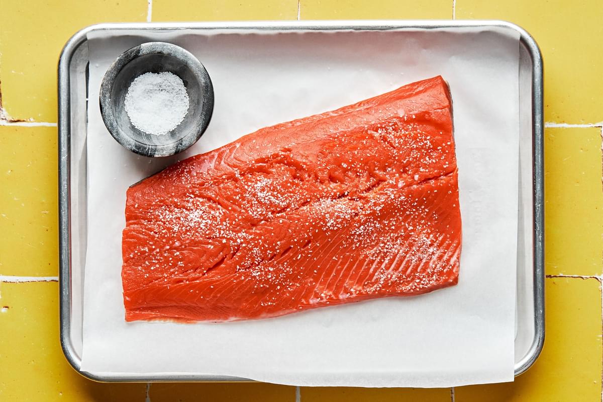 a salmon filet on a parchment lined baking sheet being seasoned with salt