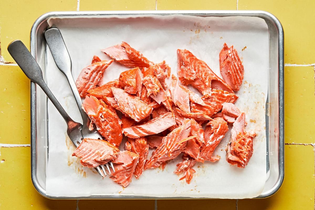 baked salmon being flaked with 2 forks on a parchment lined baking sheet