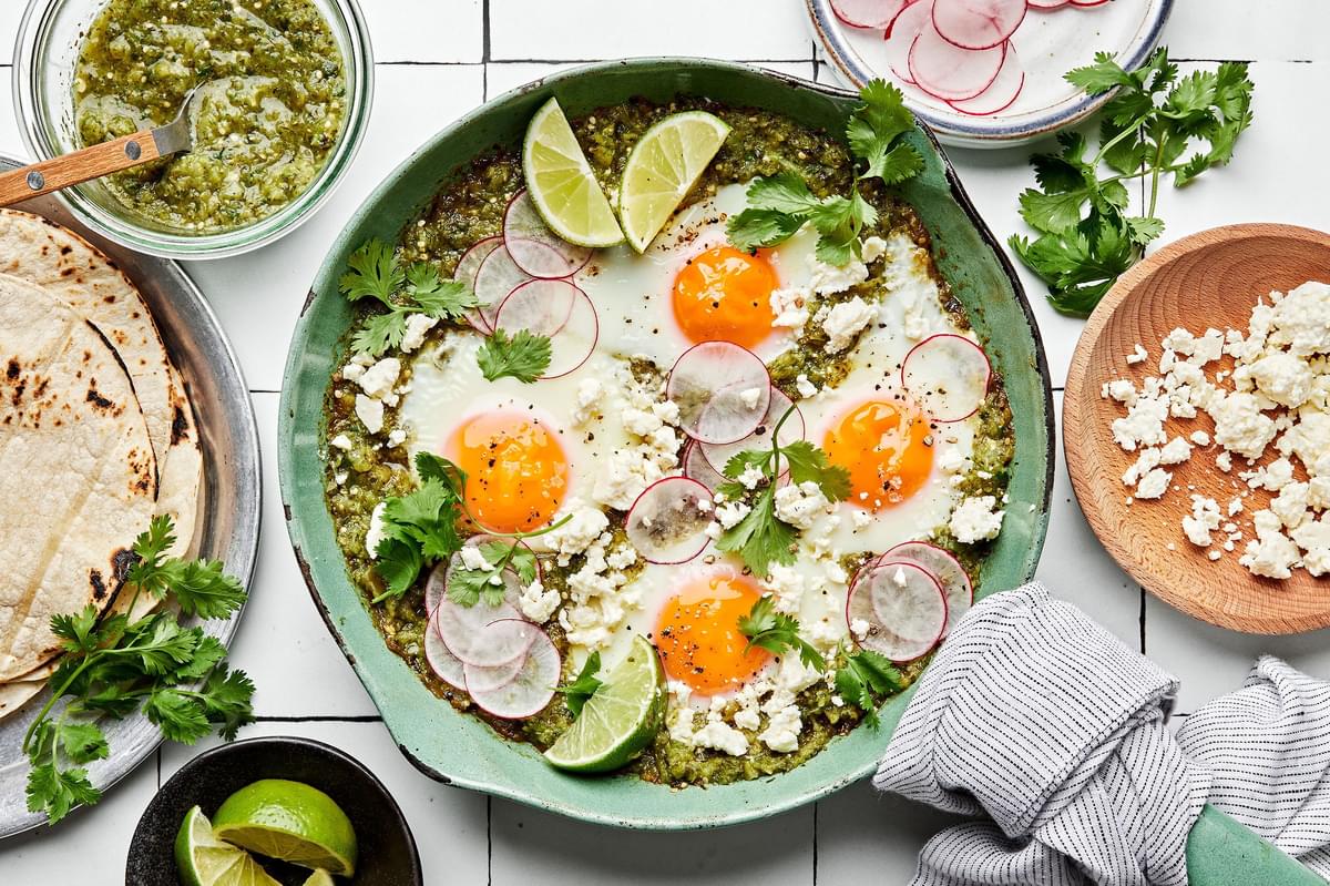 salsa verde baked eggs in a skillet sprinkled with cotija, cilantro and radishes being served with corn tortillas and limes