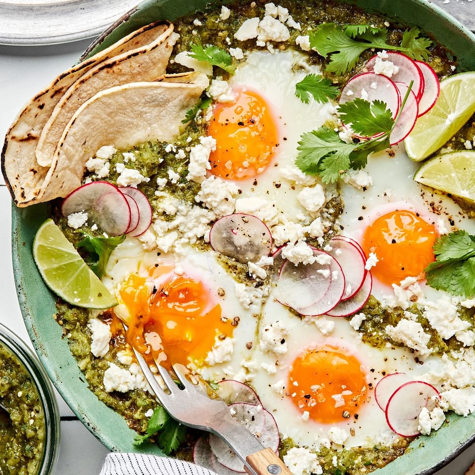 salsa verde baked eggs in a skillet sprinkled with cotija, cilantro and radishes being served with corn tortillas and limes