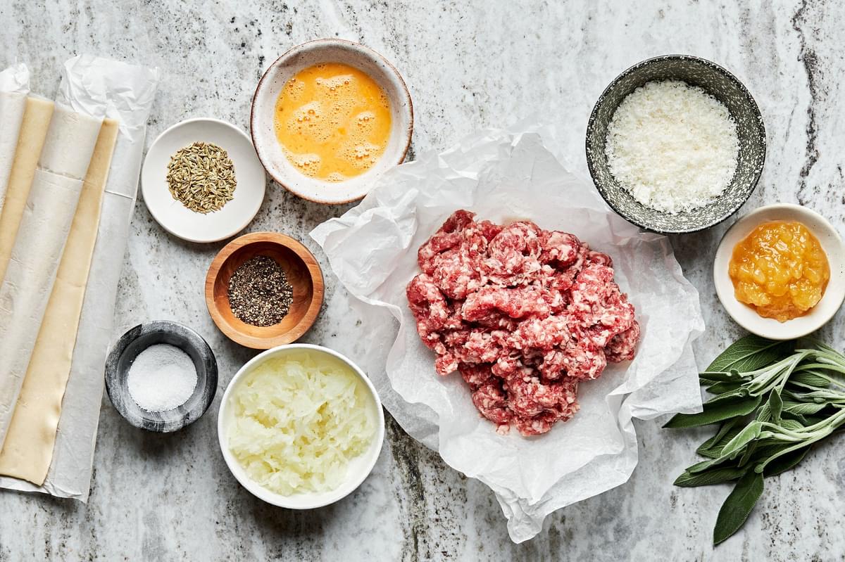ingredients for sausage rolls shown in small bowls including sausage, sage, pastry dough, parmesan cheese, eggs, breadcrumbs.