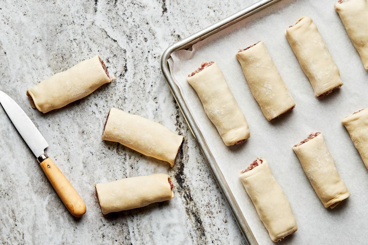 uncooked sausage rolls arranged on a baking sheet with a knife alongside