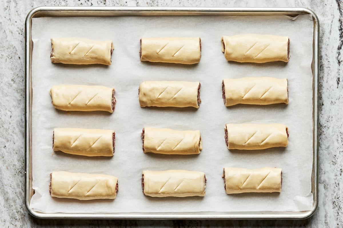 twelve uncooked sausage rolls arranged on a rimmed baking sheet and brushed with egg