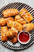 homemade sausage rolls on a black and white striped plate with a small bowl of ketchup on the plate