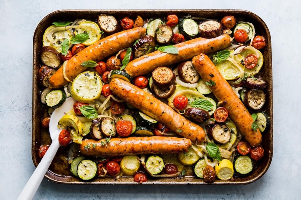 cooked sliced yellow squash, zucchini, onions and tomatoes on a sheet pan for ratatouille with sausage and a serving spoon