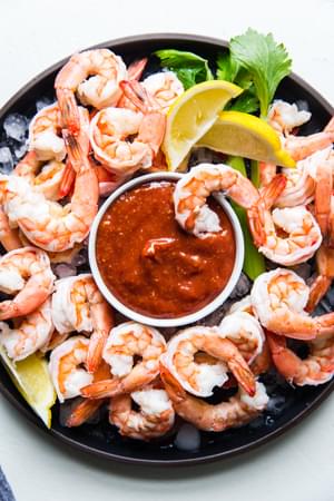 steamed shrimp on a serving platter with ice and a bowl of homemade cocktail sauce served with lemon wedges