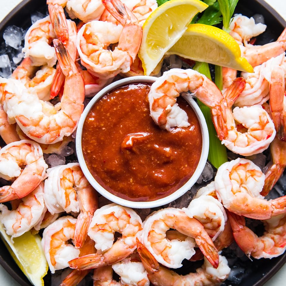 steamed shrimp on a serving platter with ice and a bowl of homemade cocktail sauce served with lemon wedges