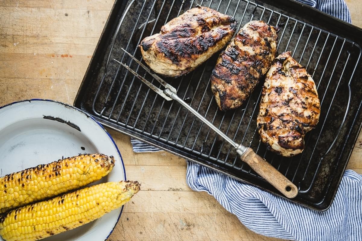 three grilled chicken breasts on a cooling rack next to a bowl of grilled corn