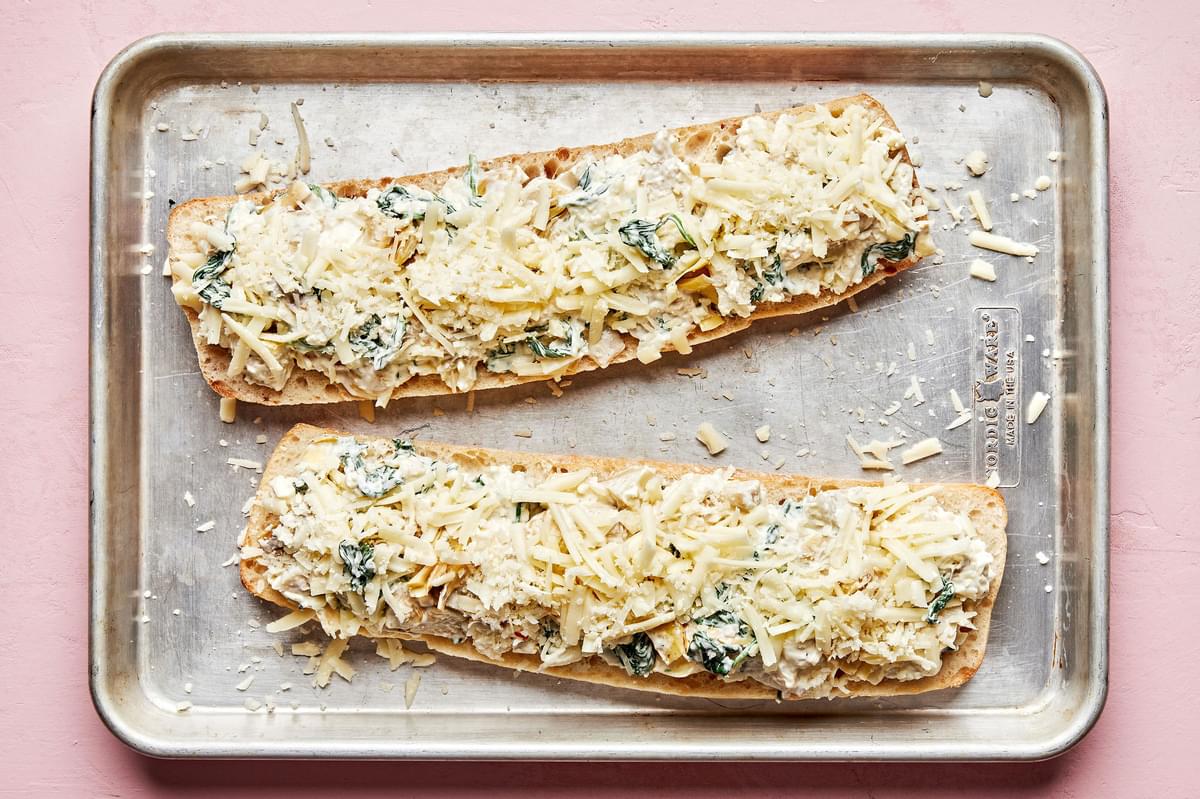 crusty bread topped with spinach, artichoke dip mixture on a sheet pan topped with mozzarella and parmesan