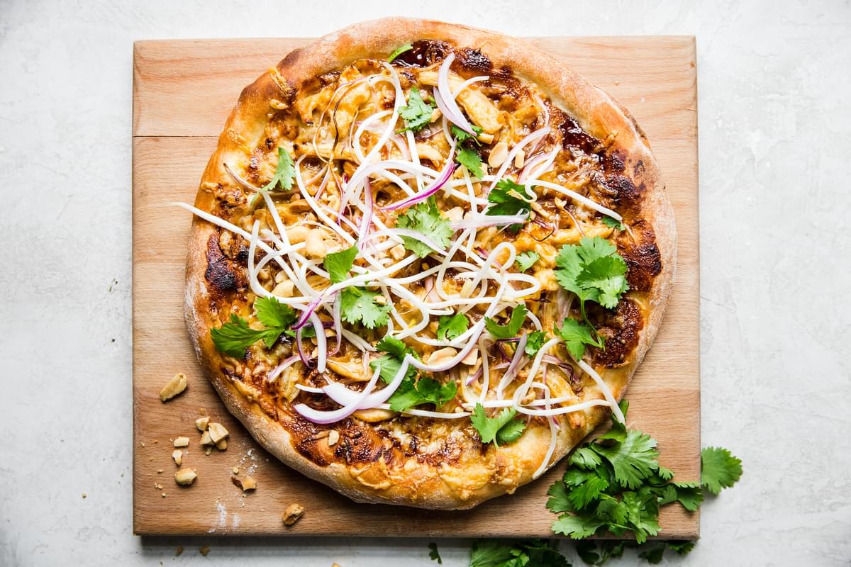 Thai style chicken pizza with cheese. onions, hoisin sauce peanuts and cilantro on a cutting board