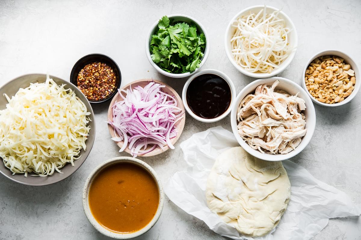 ingredients laid out for thai pizza  cheese, dough, chicken, peanut sauce, cilantro, hoisin and onions