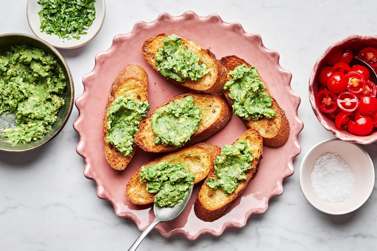 toasted baguette topped with mashed avocado surrounded by a bowl of tomato bruschetta, flaky salt and chopped basil