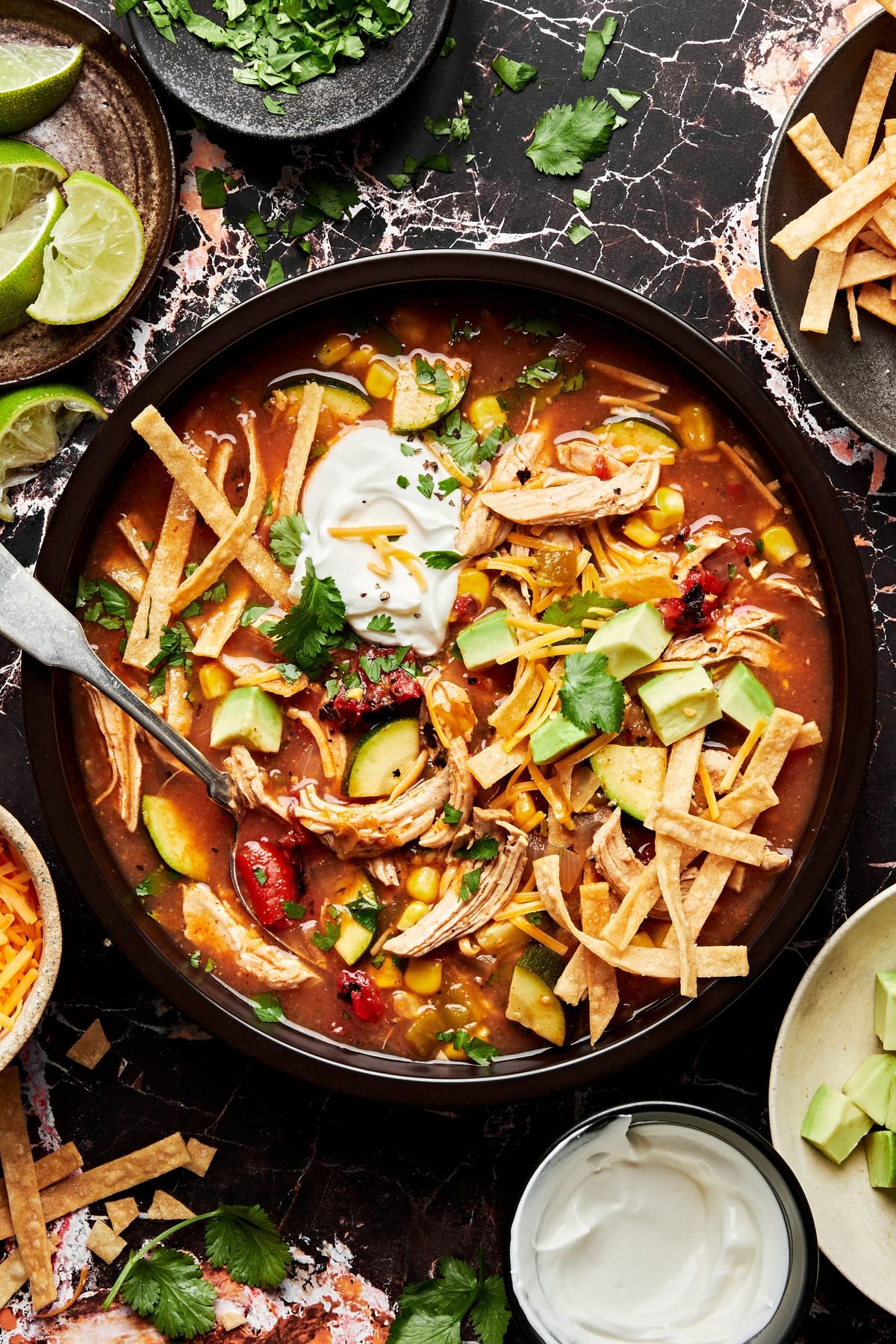 a bowl of chicken tortilla soup topped with tortilla strips, cilantro, sour cream, cheese, avocado and lime juice
