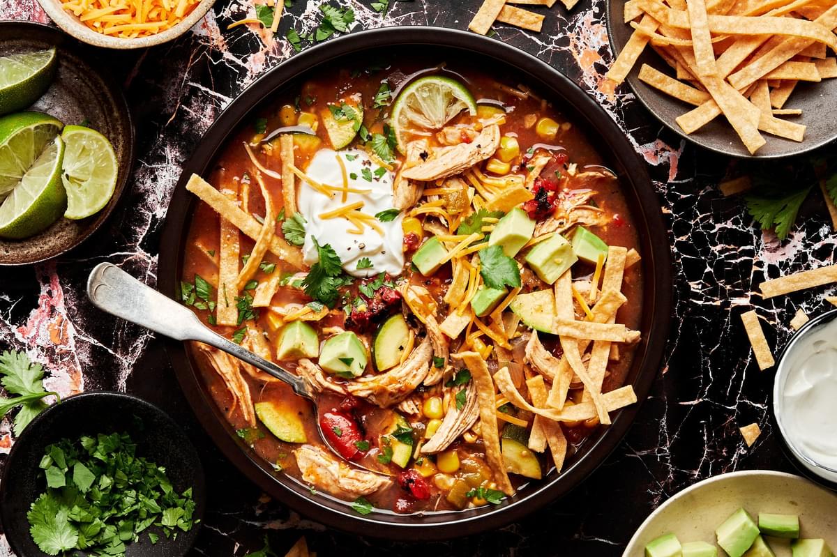 a bowl of chicken tortilla soup topped with tortilla strips, cilantro, sour cream, cheese, avocado and lime juice